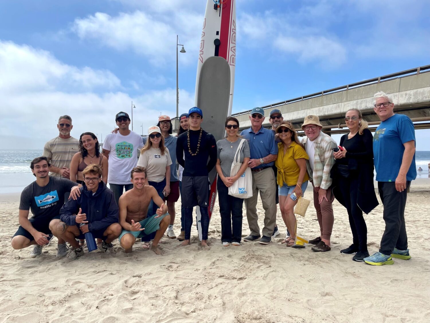 A group of people wearing Heal the Bay t-shirts pose for a photo in front of Venice Pier. In the center, Preston Lilly stands in front of his paddleboard.